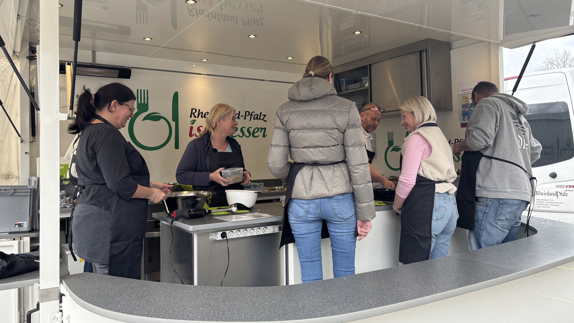 Das Bild zeigt Lehrkräfte, die im Rahmen der Veranstaltung „Der Kochbus kommt!” gemeinsam in einem aufgebauten Anhänger kochen. (2/2)