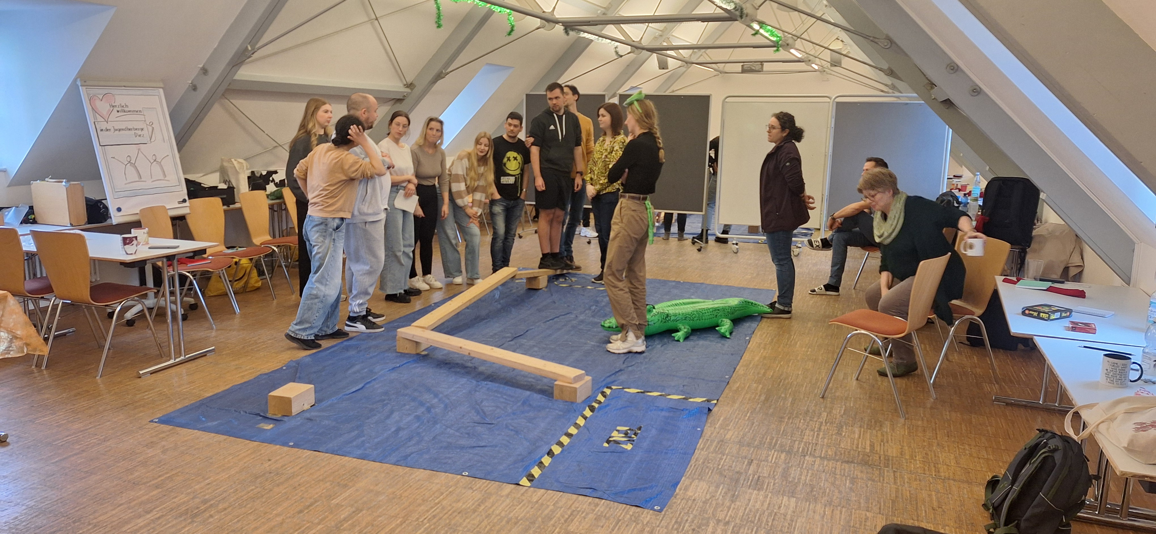 Die Gruppenmitglieder stehen um ein abgegrenztes Spielfeld, eine Person bewegt sich vorsichtig über einen Balken während die anderen Mitglieder unterstützen und anleiten.
