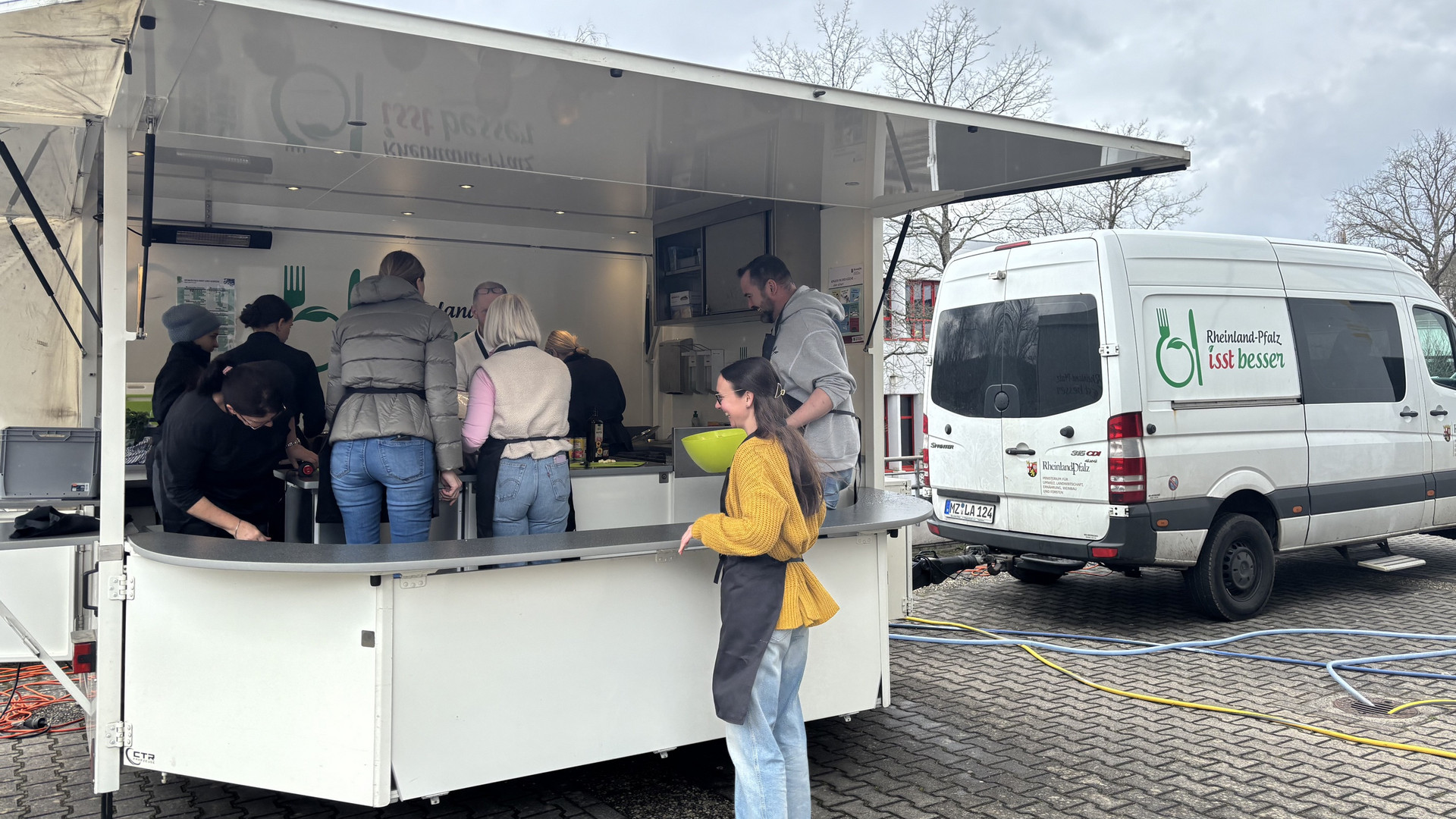 Das Bild zeigt Lehrkräfte, die im Rahmen der Veranstaltung „Der Kochbus kommt!” gemeinsam in einem aufgebauten Anhänger kochen. (1/2)