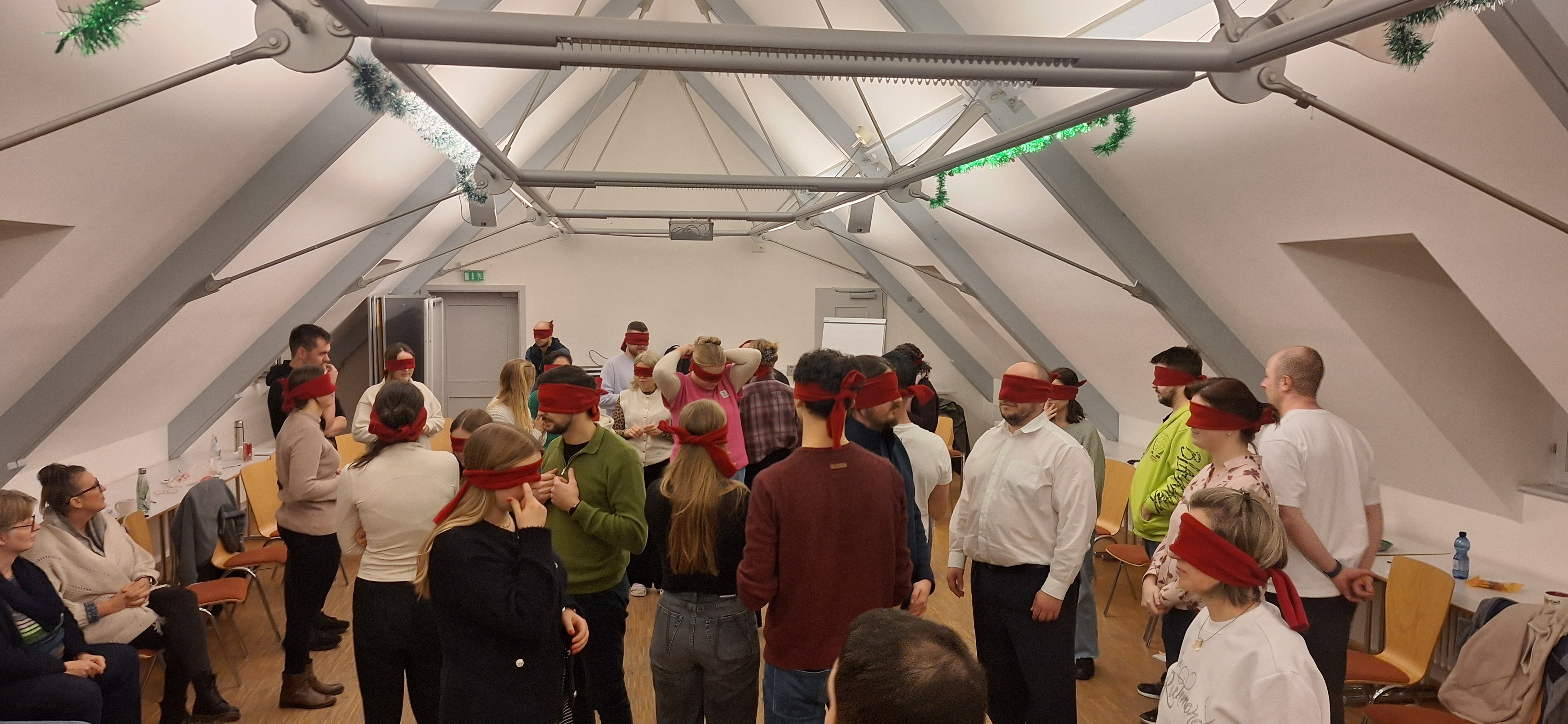 Die Gruppenmitglieder tragen Augenbinden und bewegen sich vorsichtig in einem großen Raum. Es handelt sich um eine Kommunikationsübung.