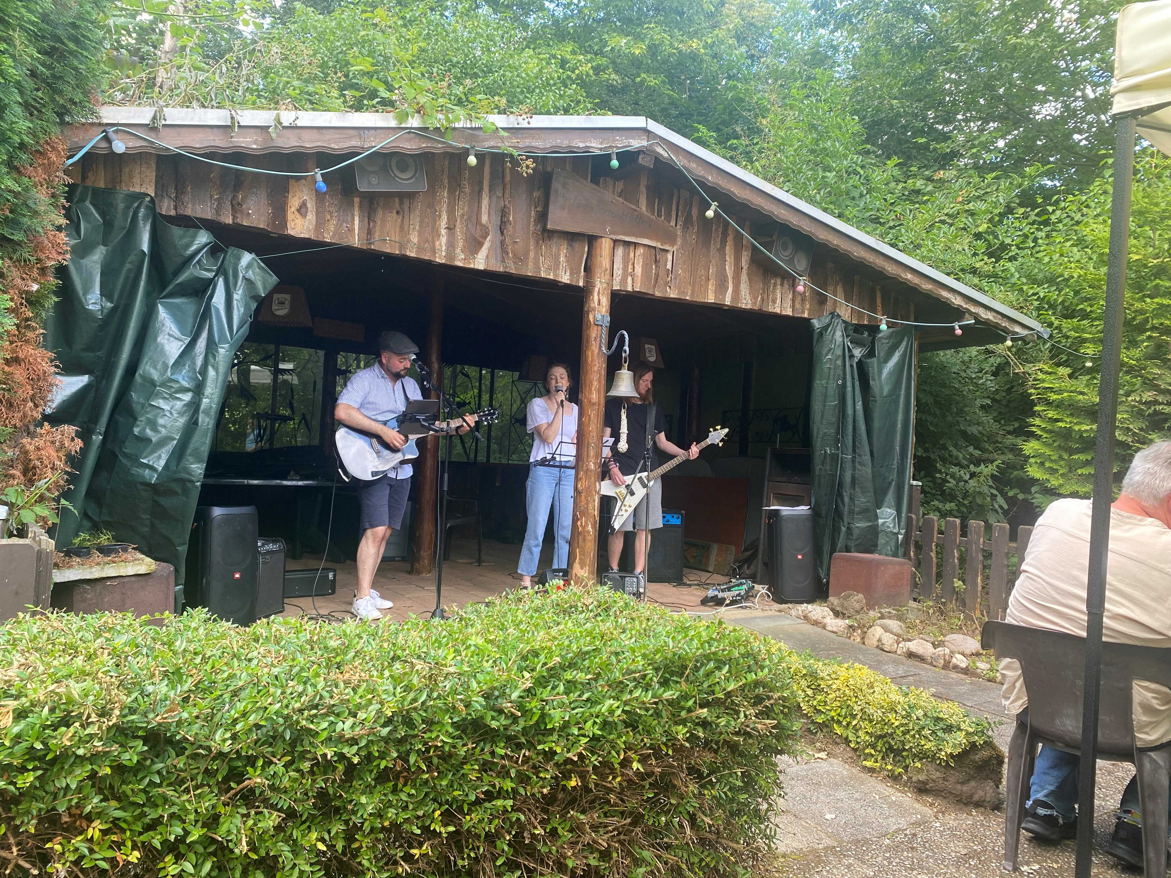 Die Band der Anwärter*innen auf der Bühne. Die Band spielt in einer als Bühne umfunktionierte Holzhütte, die unter Bäumen steht. Die Sängerin in der Mitte wird begleitet durch einen Gitarristen (links) und einen Bassisten (rechts).