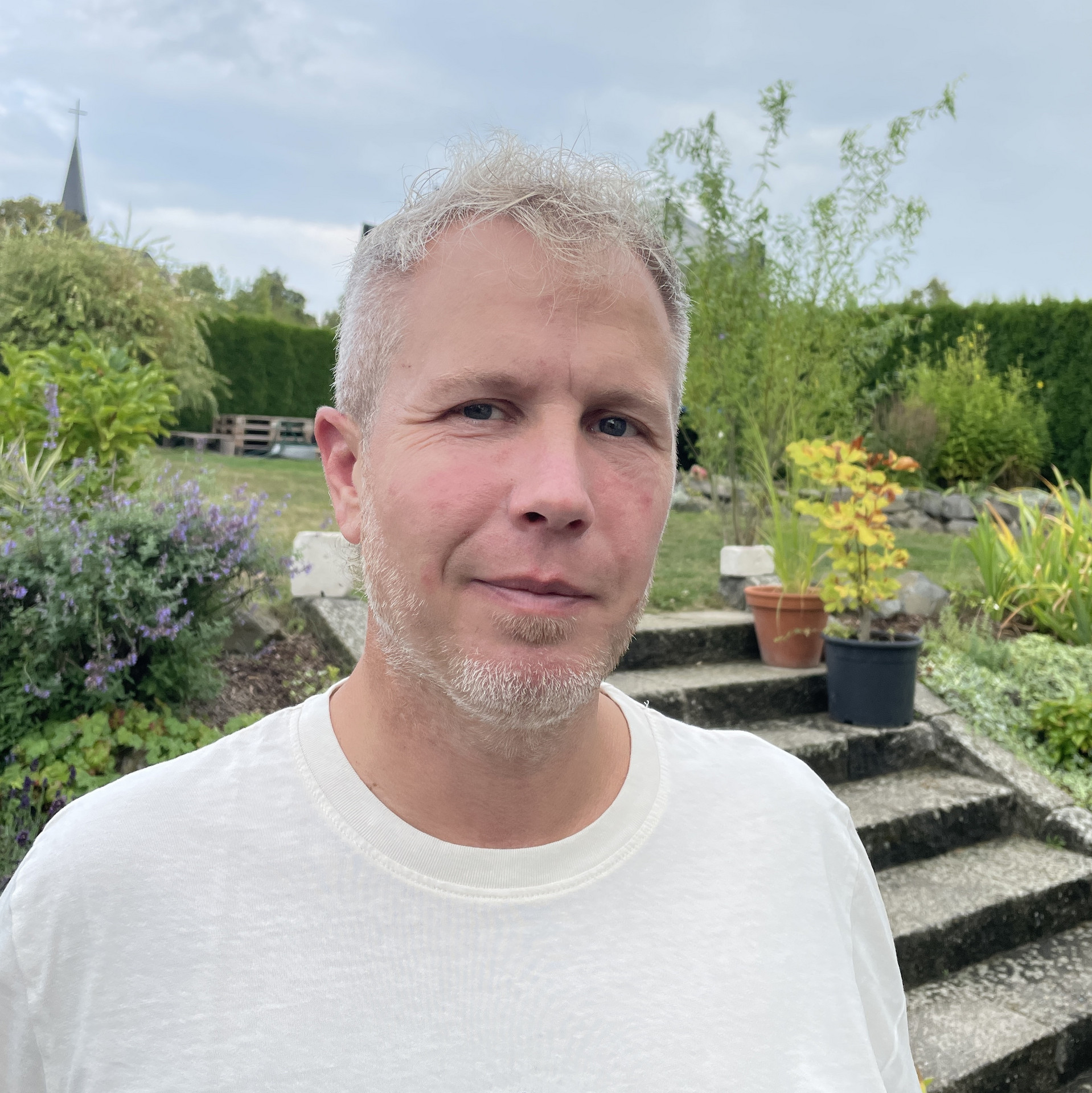 Portraitaufnahme einer Person mit kurzen hellen Haaren und weißem T-Shirt. Im Hintergrund eine Treppe, grüne Landschaft und leicht bewölkter Himmel
