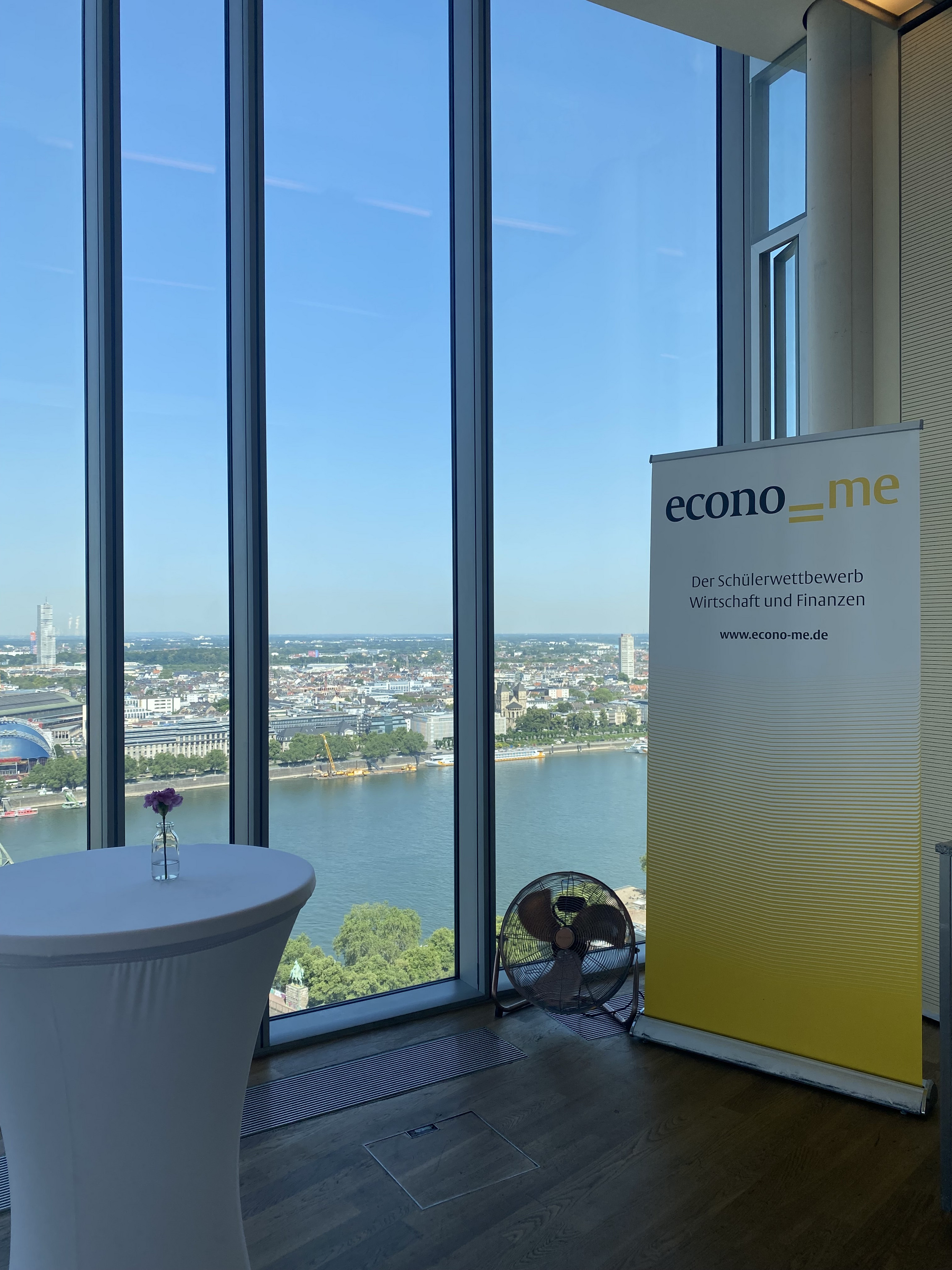 Bürogebäude der Flossbach von Storch Stiftung mit bodentiefer Fensterfront. Durch die Fensterfront ist im Hintergrund der Rhein und die Stadt Köln zu sehen. Im Vordergrund steht rechts im Raum eine Werbe-Stellwand mit dem Infotext "econo=me - Schülerwettbewerb für Finanzen" und links ein weiß eingedeckter Gesprächstisch.