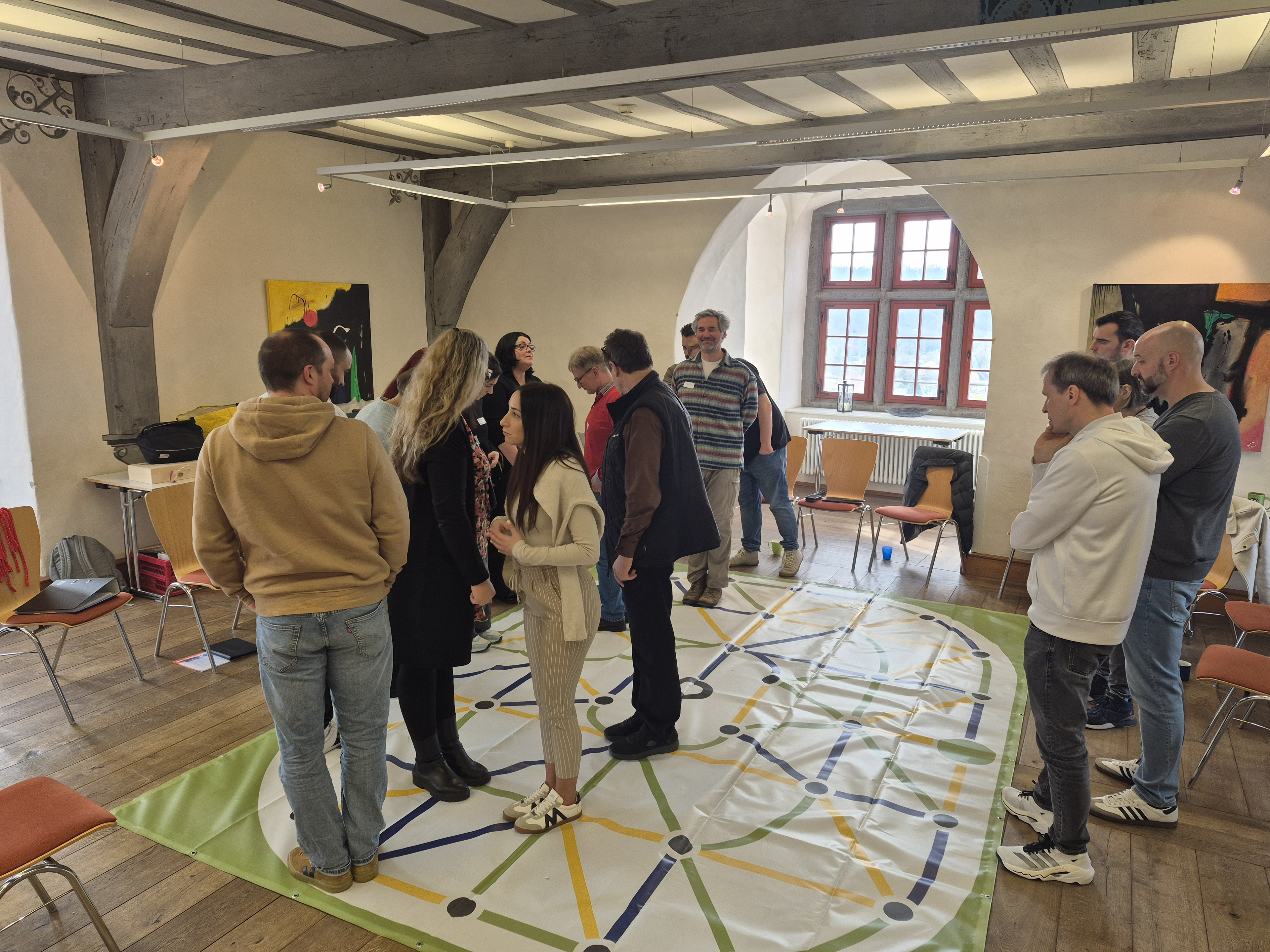 In einem Seminarraum der Jugendherberge Diez versuchen 14 Teilnehmer/-innen den richtigen Weg vom Start zum Ziel auf einer Plane mit verschiedenfarbigen Wegabschnitten zu finden (Tool: Corridor of Colors).