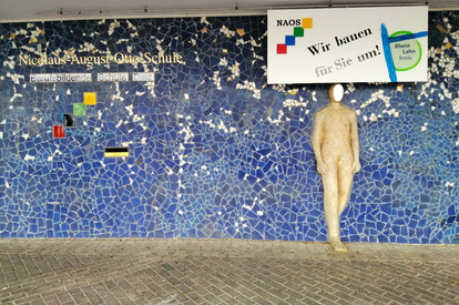 Kunstwerk aus blauem Mosaik an der Erdgeschossfassade des Haupteingangs der BBS Nicolaus-August-Otto Schule in Diez. Davor befindet sich eine lebensgroße Betongussfigur einer Person, deren Gesicht durch ein Spiegel ersetzt ist. An der Wand ist ebenfalls ein gestaltetes Baustellenschild der Klasse der Maler und Lackierer des Ausbildungsjahres 2019, welches das Logo der NAOS und des Rheinlahn Kreises trägt mit der Aufschrift "Wir bauen für Sie um!".  damit wird auf seit 2019 laufende Bauarbeiten an der Schule hingewiesen.