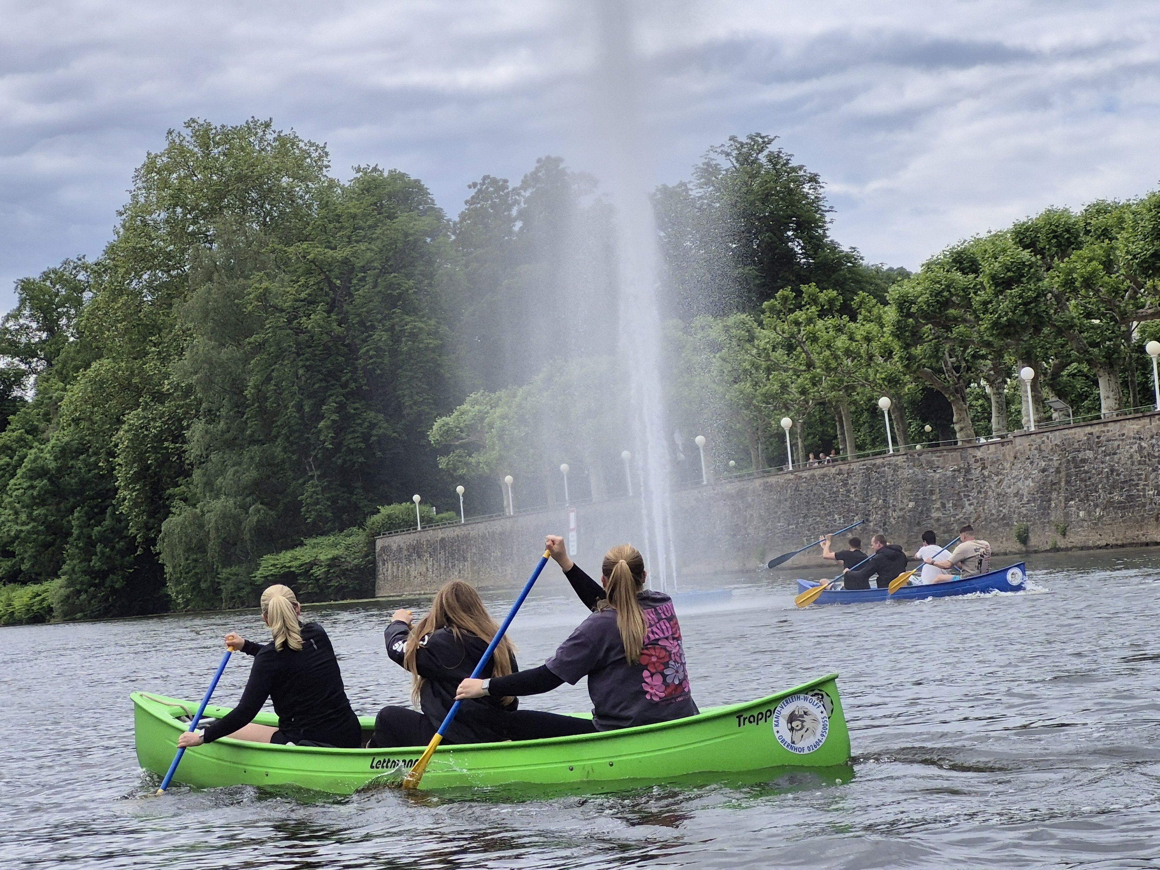 Auf der Lahn in Bad Ems: Im Hintergrund sind große grüne Bäume und der blaue Himmel mit vielen weißen Wolken zu sehen. Davor sieht man das steile Steinufer der Fußgängerpromenade mit Lampen, davor ein Springbrunnen mit einer mehrere Meter hohen Fontäne. Rechts davon fährt ein Kanadier mit vier Männern in diese Fontäne rein. Links davon im Vordergrund fährt ein Kanadier mit drei Frauen, die das Männerboot beobachten.