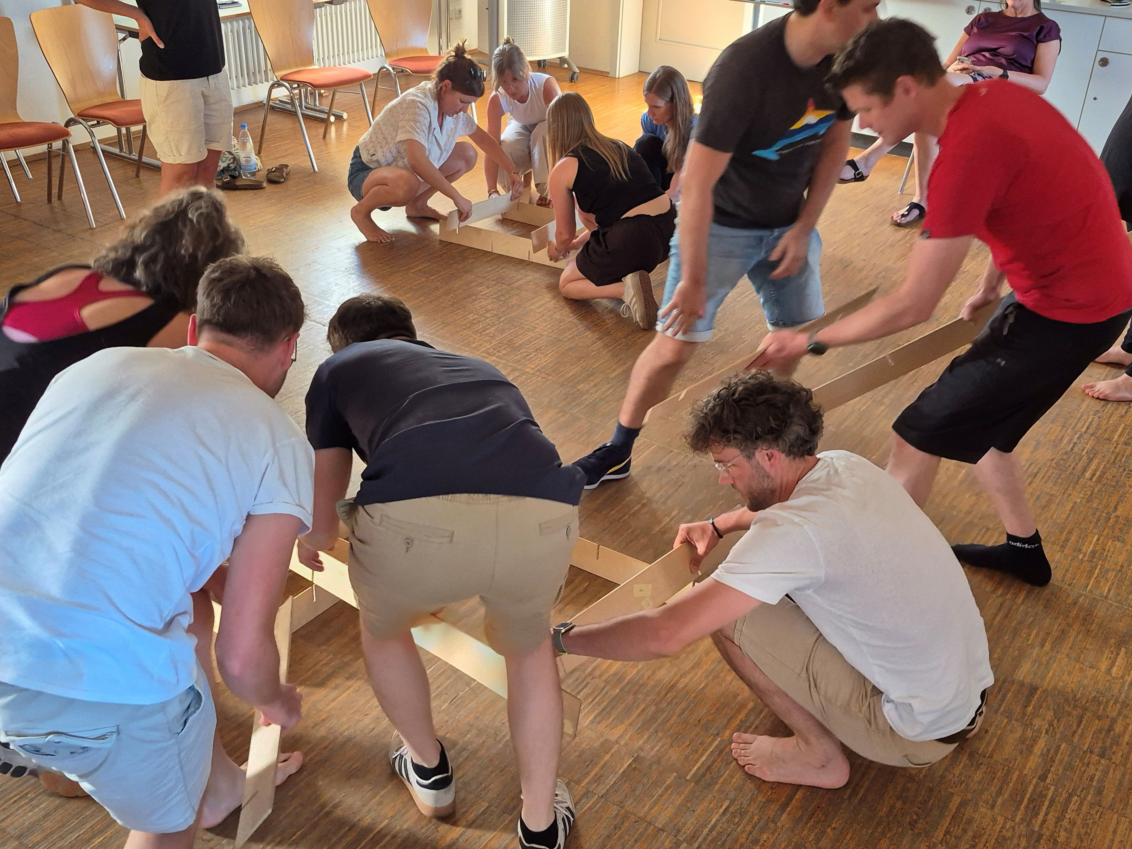 In einem Seminarraum sind elf Anwärterinnen beim Tool Heckmeck in Aktion, wobei alle versuchen, jeweils ein Ein-Meter langes Holzteil zu einer vorgegebenen Konstruktion möglichst schnell richtig auf einem Holzboden zusammenzustecken. Im Vordergrund sieht man bückend links eine Anwärterin und rechts davon fünf Anwärter. Dahinter sind vier weitere Anwärterinnen bückend bzw. knieend mit ihren Holzteilen zu sehen. Links davon steht ein Anwärter (der Kopf ist nicht zu erkennen), im Hintergrund beobachtet sitzend die Fachleiterin, die das Tool angeleitet hat, das Geschehen (auch hier ist das Gesicht nicht zu erkennen). Links am Rand hinter dem stehenden Anwärter stehen vier Stühle.