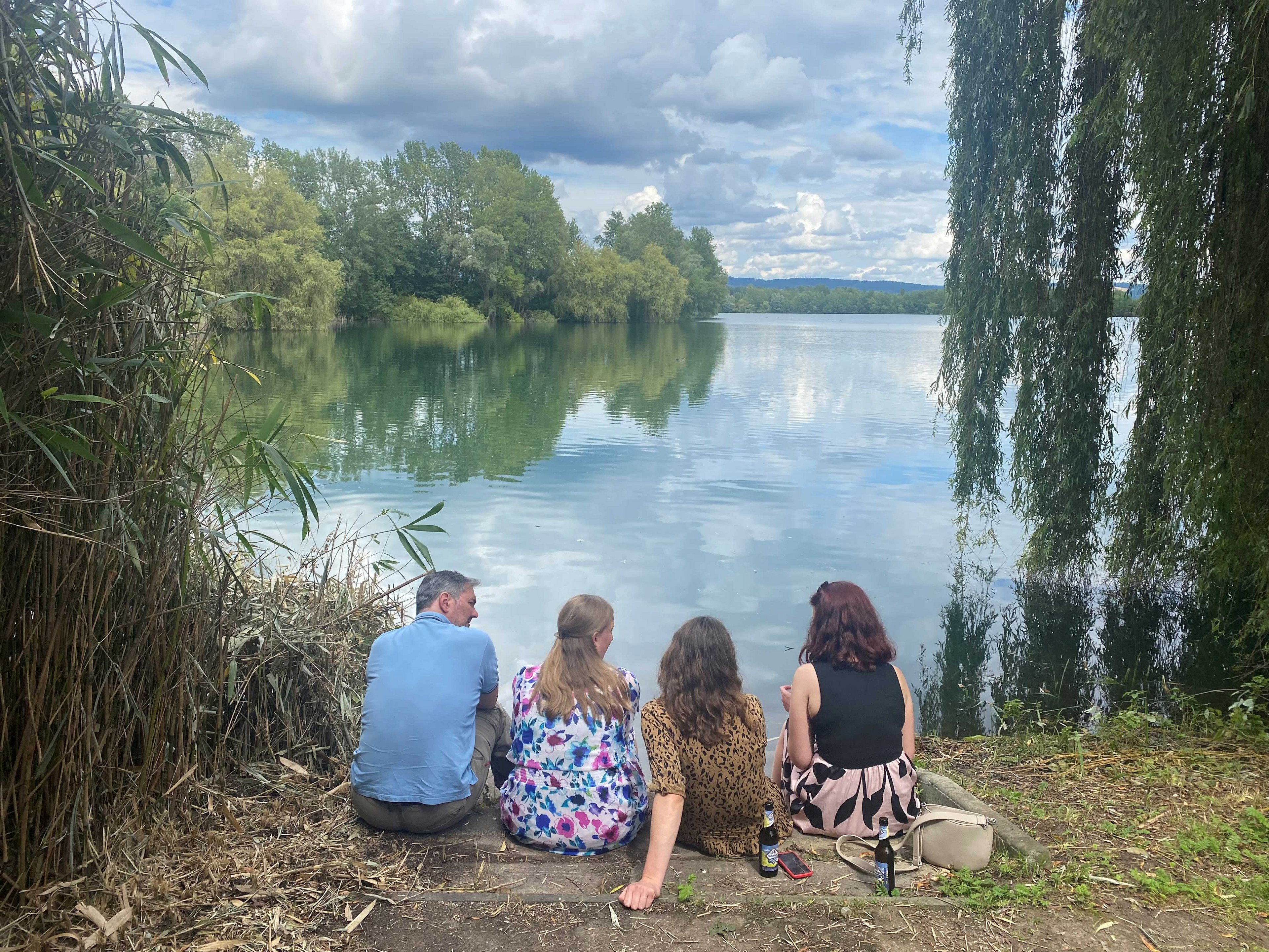 Eine Gruppe von vier Personen sitzt am Ufer des Steinsees mit dem Rücken zur Kamera. Ein Fachleiter unterhält sich angeregt mit drei Anwärterinnen. Das Bild wirkt mit den sich im Wasser spiegelnden Wolken und den bis ins Wasser reichende Äste der Trauerweiden malerisch.