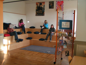Aufnahme einer Leseecke: sonniger, heller Eckraum; Holz-Sitzstufen mit Kissen, darauf vier Mädchen, in die Lektüre von Büchern vertieft.