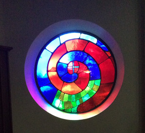 Fenster in der Hundertwasser-Kirche St. Barbara, Bärnbach/Steiermark: Bleiglas mit Segmenten in verschiedenen Farben. Zwei ineinander verlaufende Spiralen, von denen die eine in rötlichen Tönen gehalten ist, die andere blaue und grüne Elemente enthält.