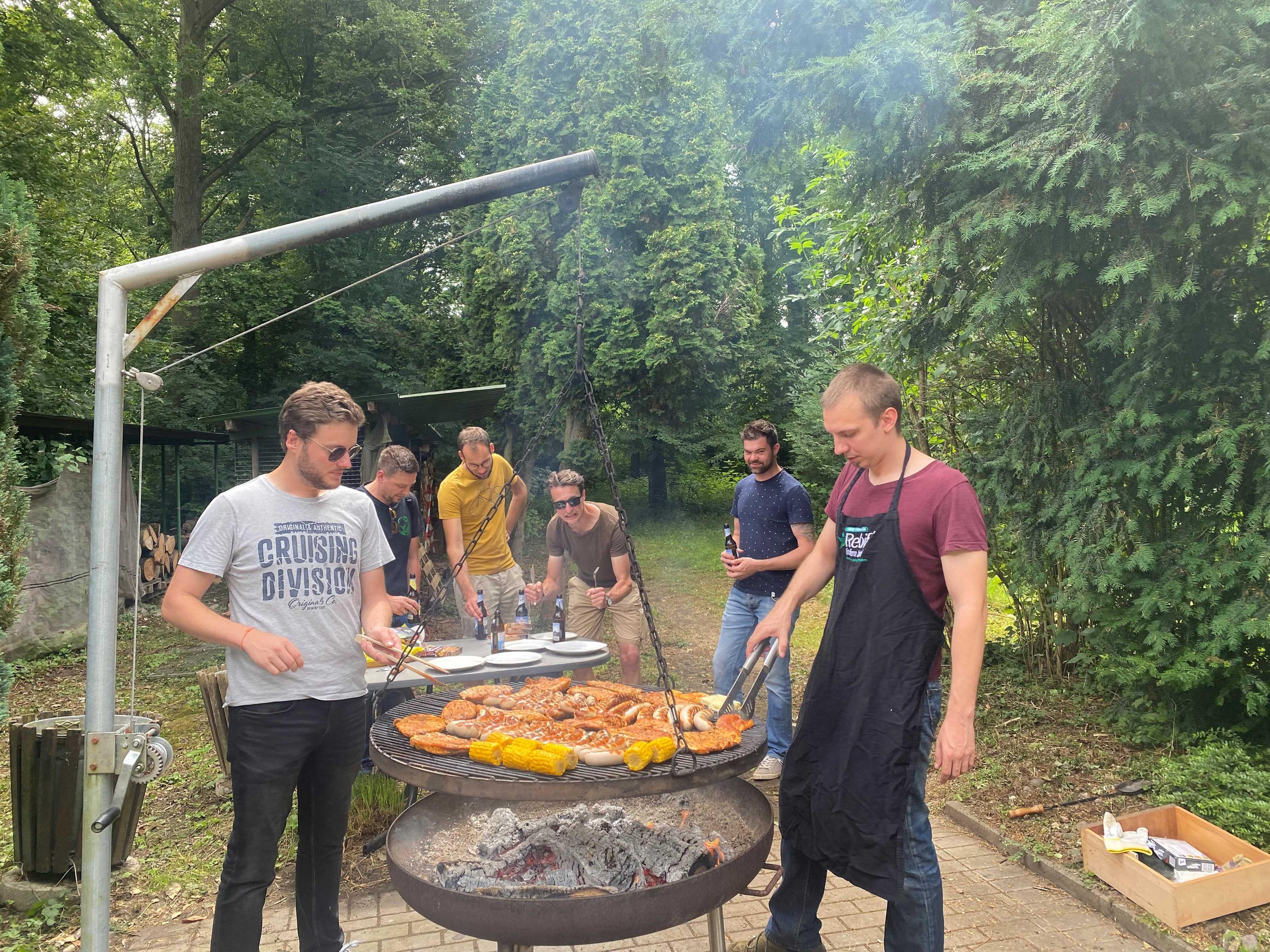 Das Grillteam am großen Schwenkgrill. Sechs junge Männer kümmern sich um Grill und Grillgut. Auf dem Grill liegen Würstchen, Steaks, Meiskolben und Grillkäse.