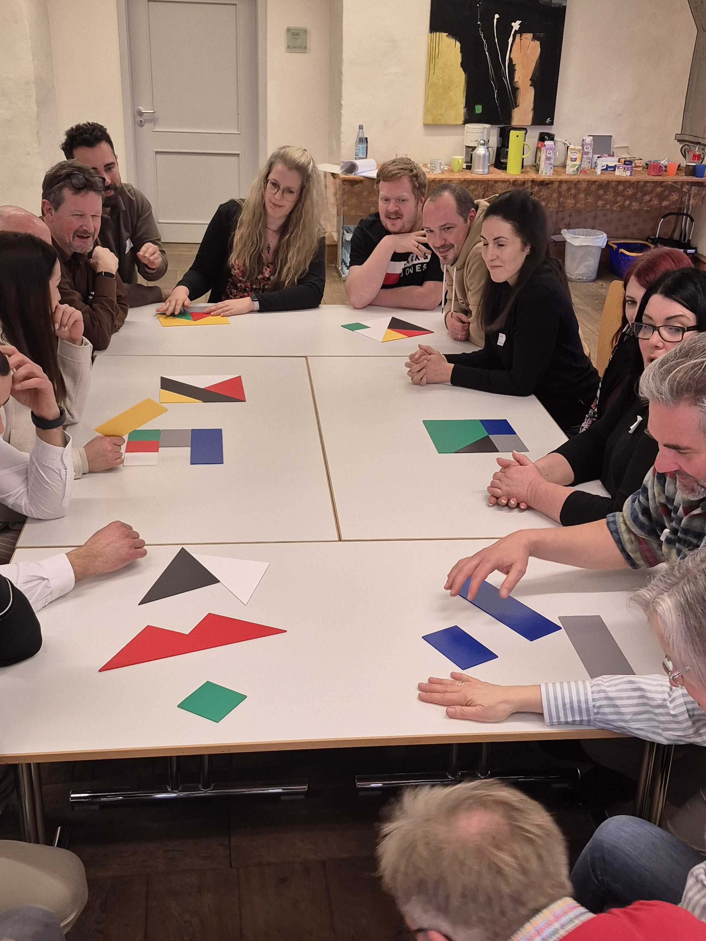 15 Teilnehmer/-innen sitzen um einen großen Tisch, auf dem verschiedenfarbige Puzzleteile liegen. Ziel ist es, 7 Quadrate so zu legen, dass sich jeweils keine Farbe im Quadrat wiederholt.