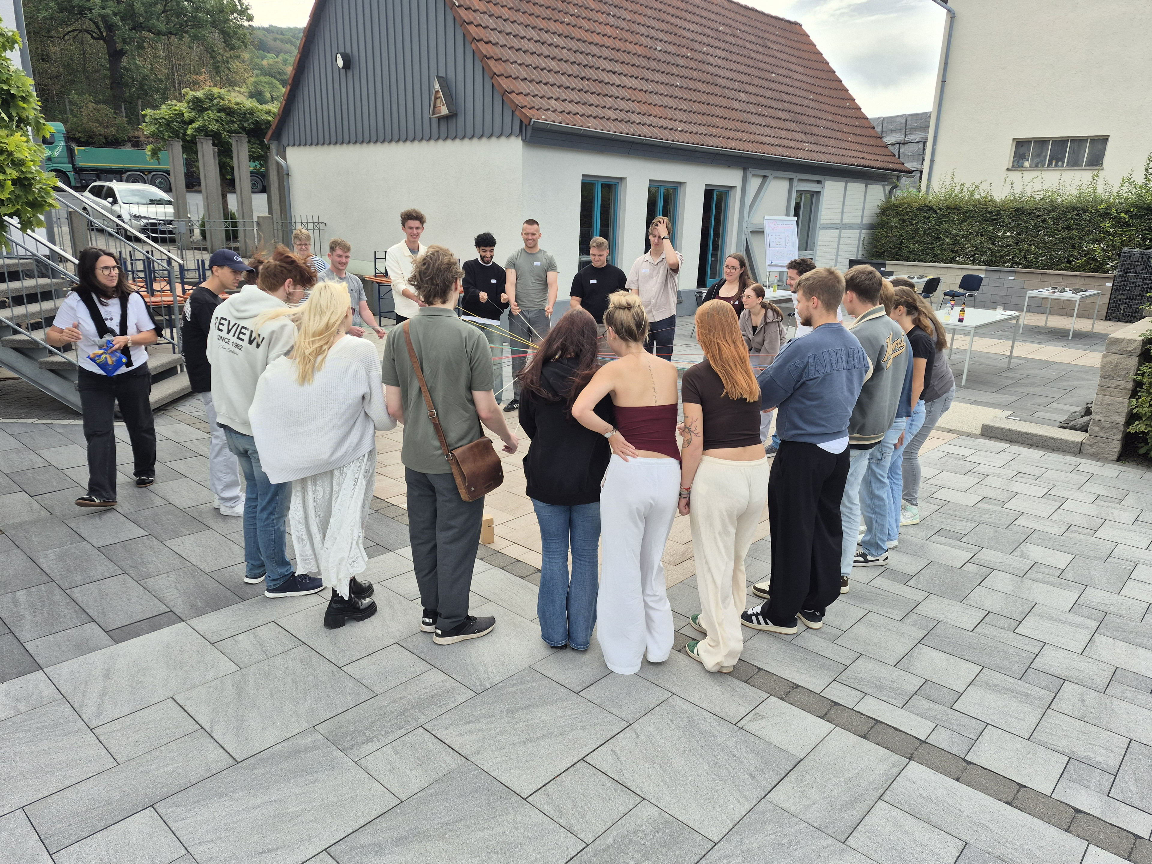 23 Auszubildende stehen auf einem Hof mit verschiedenen Steinplatten (Musterausstellung des Betonwerks Seiferts vom Unternehmen FCN) im Kreis und halten jeweils ein bis 2 Schnüre in der Hand, mit denen sie einen Kran bewegen, um 8 Holzklötze übereinander zu stellen. Im Hintergrund ist in der Mitte ein Gebäude mit 2 Seminarräumen zu sehen sowie rechts eine Hecke und links Bäume.