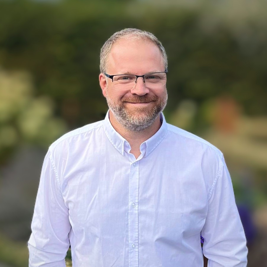 Aufnahme einer Person in hellem Hemd mit hellen Haaren und hellem Bart sowie einer Brille. Im Hintergrund verschwommen die Konturen von grünen Pflanzen