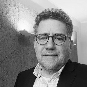 Schwarz-weiß Portrait einer Person mit Brille und kurzen Haaren. Die Person trägt ein helles Hemd und ein dunkles Jackett.