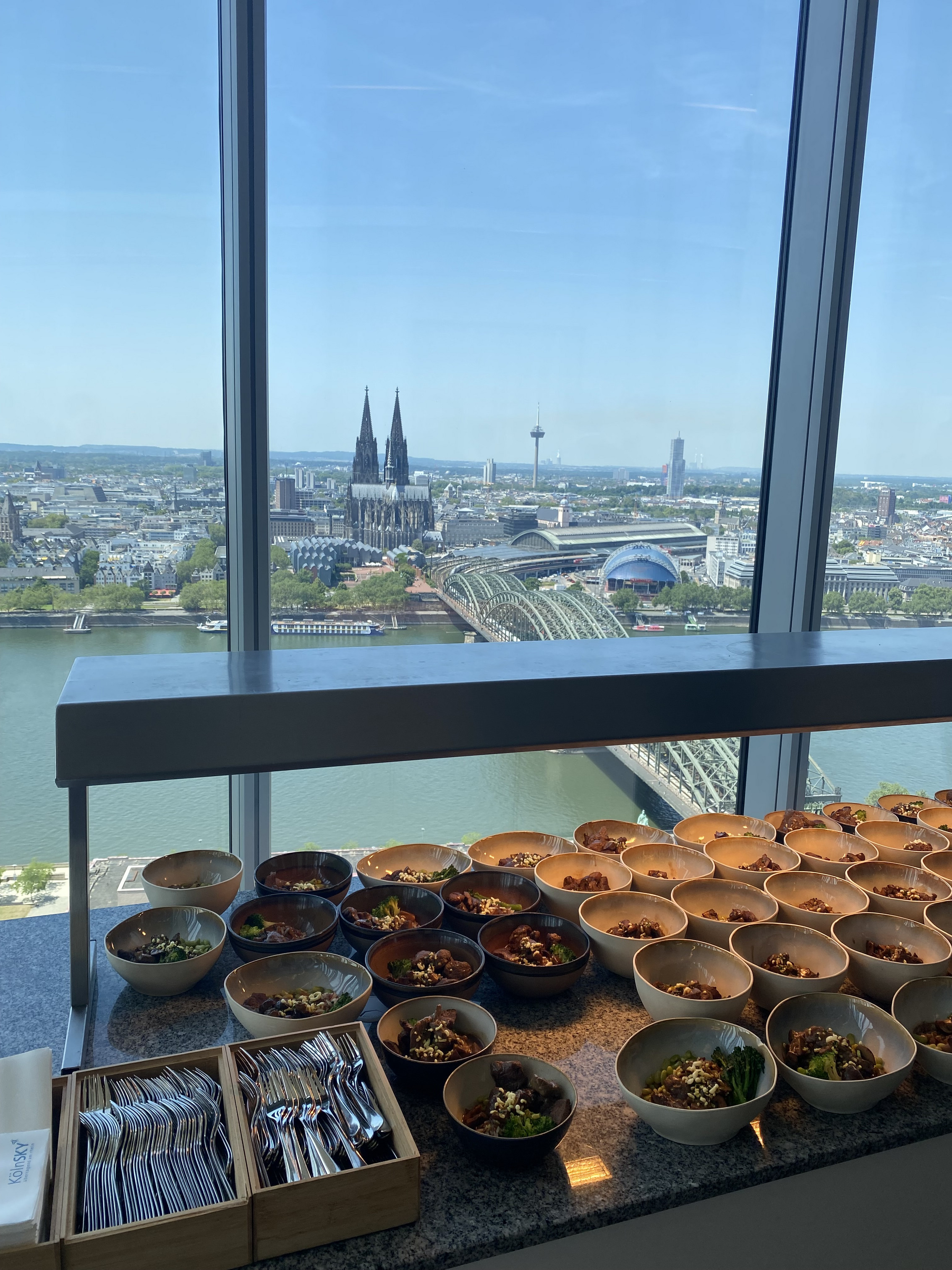 Bürogebäude der Flossbach von Storch Stiftung mit bodentiefer Fensterfront. Durch die Fensterfront ist im Hintergrund der Rhein, die Stadt Köln, der Kölner Dom und der Hauptbahnhof zu sehen. Im Vordergrund steht ein aufgebautes Buffet.