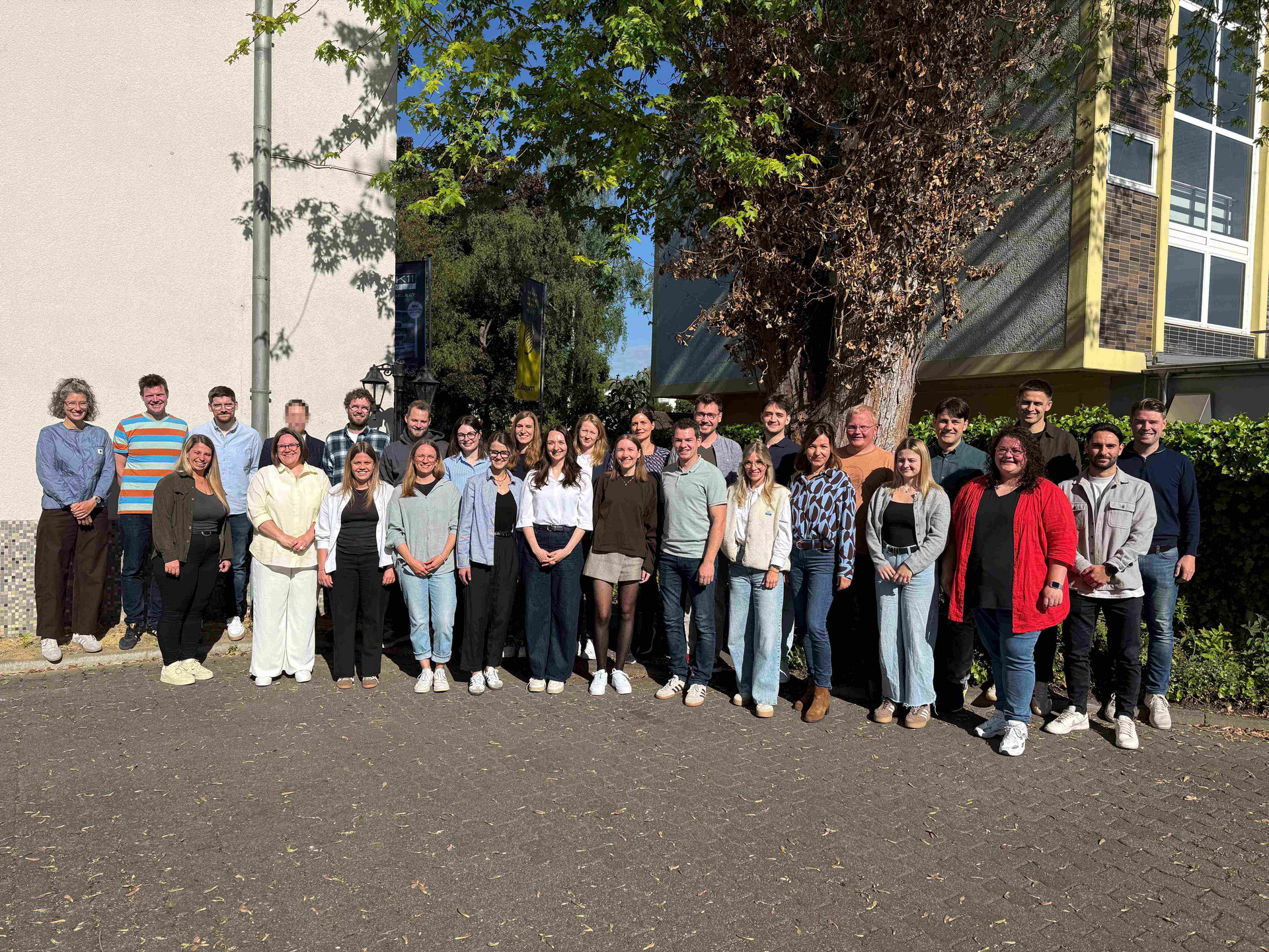 Die Ausbildungsgruppe F26 hat sich für das Foto in zwei Reihen aufgestellt. Zu sehen sind 29 Personen, die vor einem Baum im Hof des Seminargebäudes stehen.