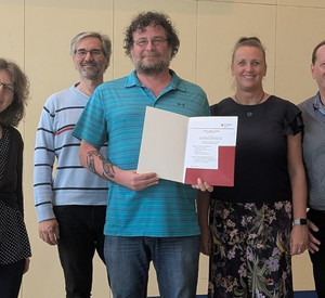 Gruppenbild mit 5 Personen. Die mittlere Person hält eine geöffnete Mappe mit einer Urkunde darin.