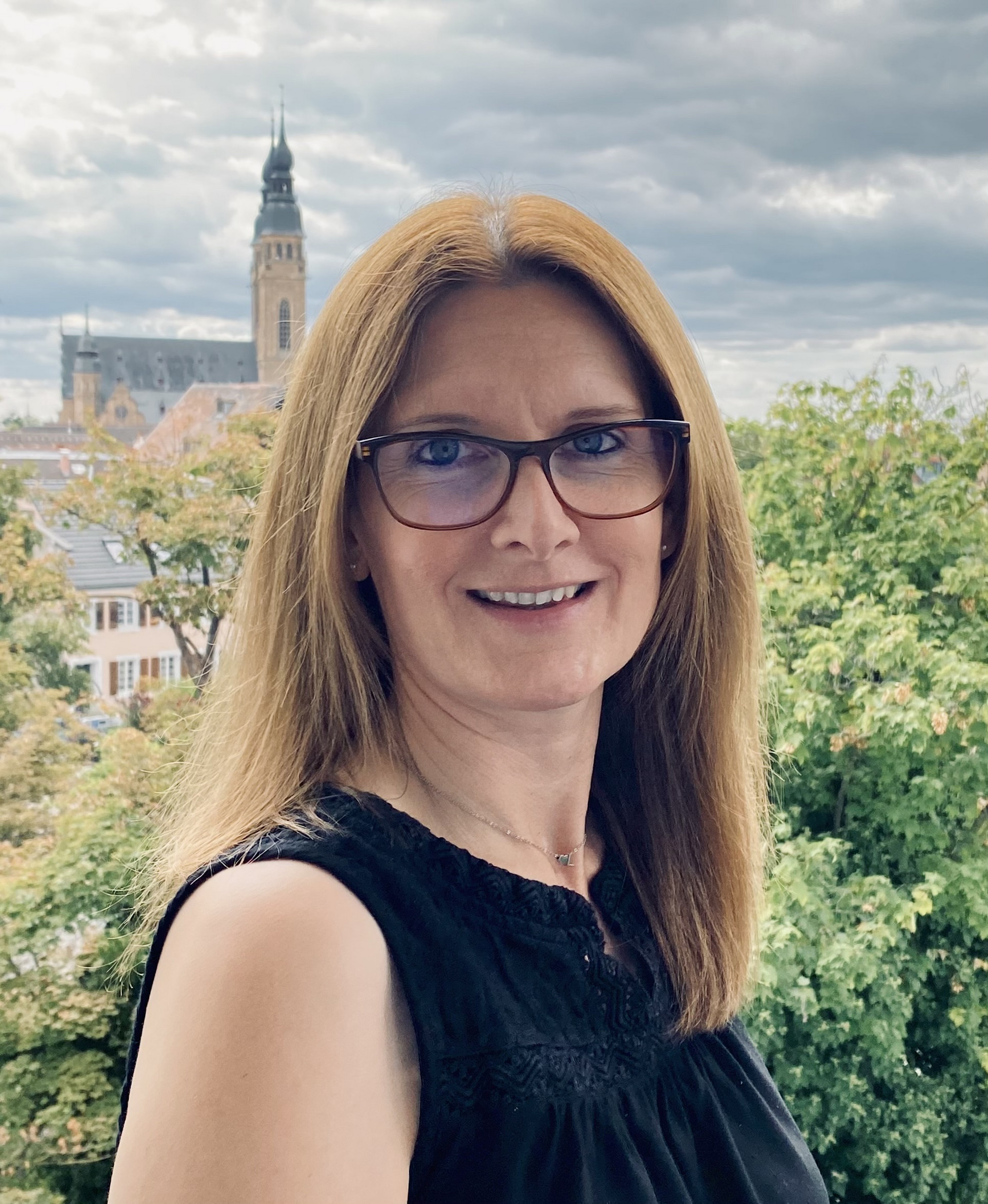 Portraitaufnahme einer Person mit langen Haaren und Brille, dunkles ärmelfreies Oberteil. Im Hintergrund Bäume, bewölkter Himmel sowie Gebäude