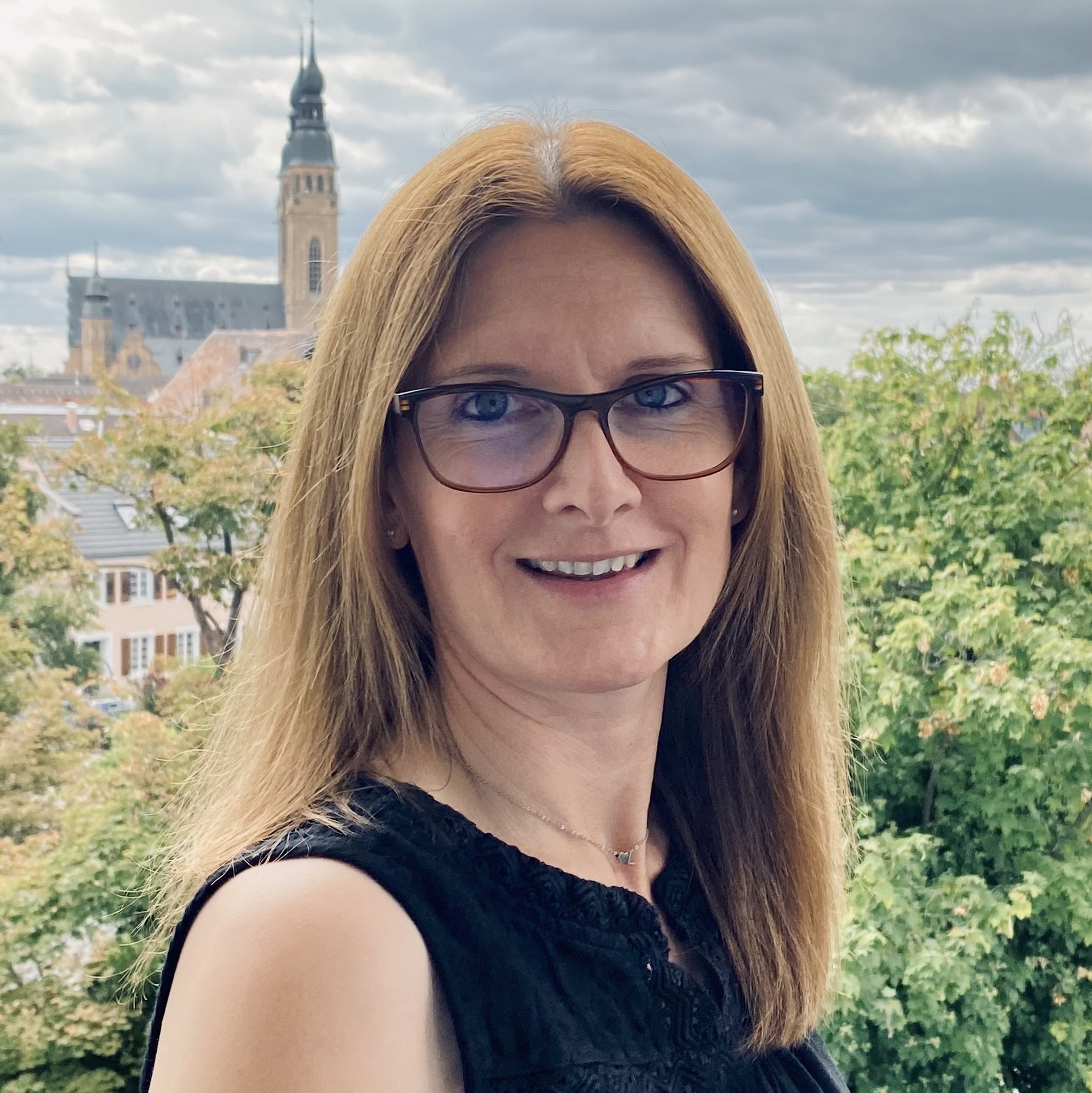 Portraitaufnahme einer Person mit langen Haaren und Brille, dunkles ärmelfreies Oberteil. Im Hintergrund Bäume, bewölkter Himmel sowie Gebäude