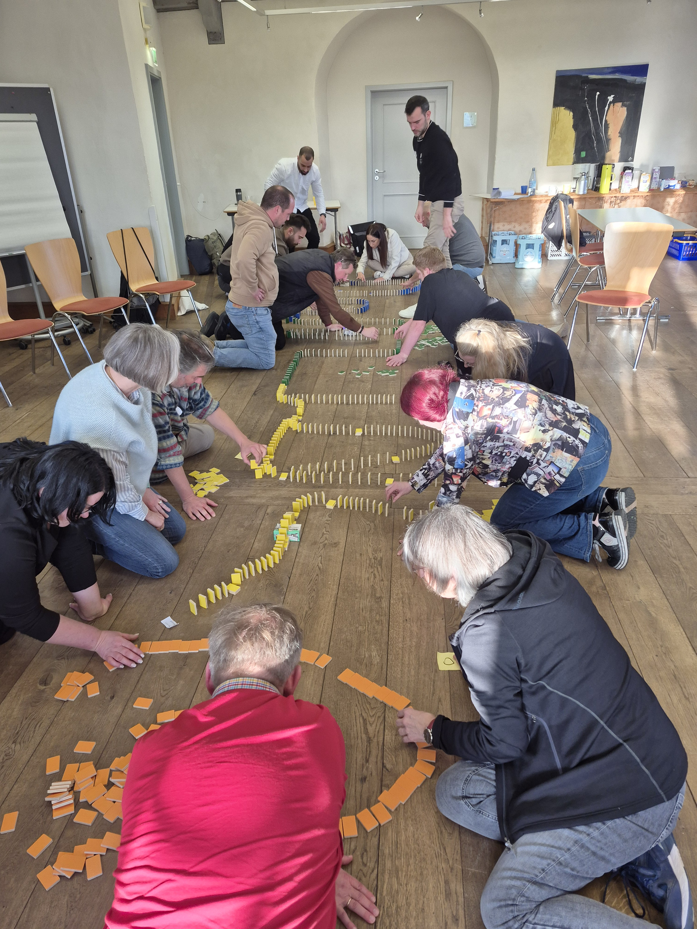 Auf dem Fußboden eines Seminarraumes knieend stellen 14 Teilnehmer/-innen in 4 Gruppen Dominosteine so auf, dass sie später durch ein einmaliges Anstoßen alle umfallen. Dabei soll das Wort Domino dargestellt sowie 4 Hindernisse eingebaut werden.