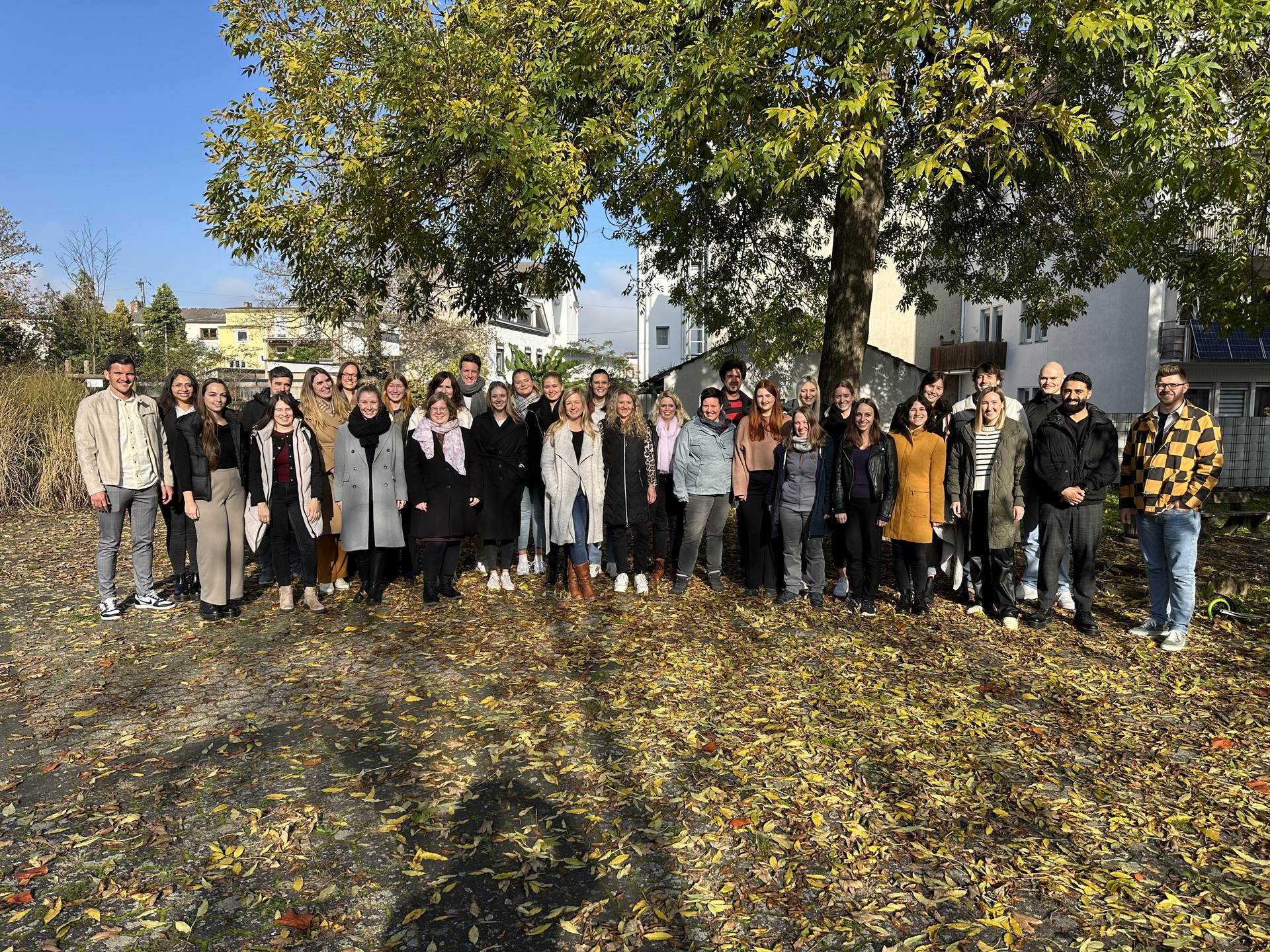 Gruppenfoto mit den Anwärter*innen der Stammgruppe H24 des Studienseminars für das Lehramt an berufsbildenden Schulen in Neuwied. Die Gruppe steht gut gelaunt an einem herbstlichen Tag unter einem Baum, dessen Laub den Boden bedeckt. Im Hintergrund sind noch Häuser zu erkennen.