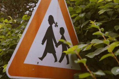 Das Bild zeigt ein Verkehrsschild. Das Schild ist dreieckig mit einem orangefarbenen Rand.     Darauf sind eine Frau und ein Kind abgebildet, die Hand in Hand gehen. Der Hintergrund ist weiß.