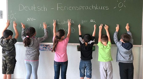 Kinder stehen mit dem Gesicht zu, und den Händen nach oben gestreckt vor einer Tafel. Auf der Tafel steht "Deutsch Feriensprachkurs".