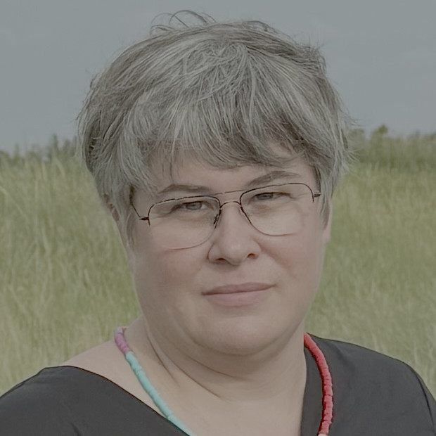Portrait einer Person mit kurzen Haaren und Brille mit Kette und dunklem Oberteil. Im Hintergrund Himmel und grüne Landschaft