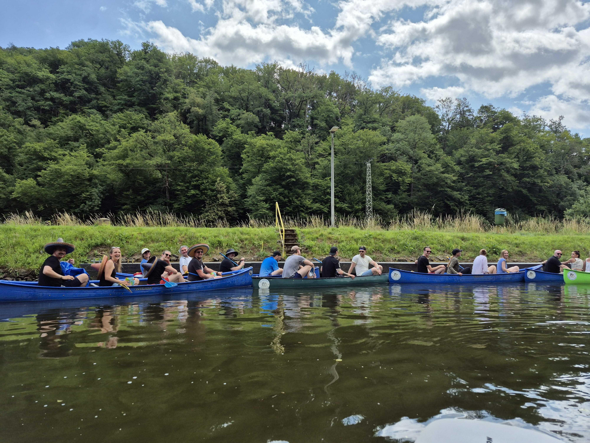 Vor der ersten Schleuse Hollerich warten 6 blaue Kanus mit 20 Anwärterinnen und Anwärtern an einer Anlegestelle. Im Vordergrund ist die Lahn zu sehen, im Hintergrund erst Wiese und dahinter zahlreiche Laubbäume. Am oberen Bildrand sieht man den Himmel mit weißen Wolken.