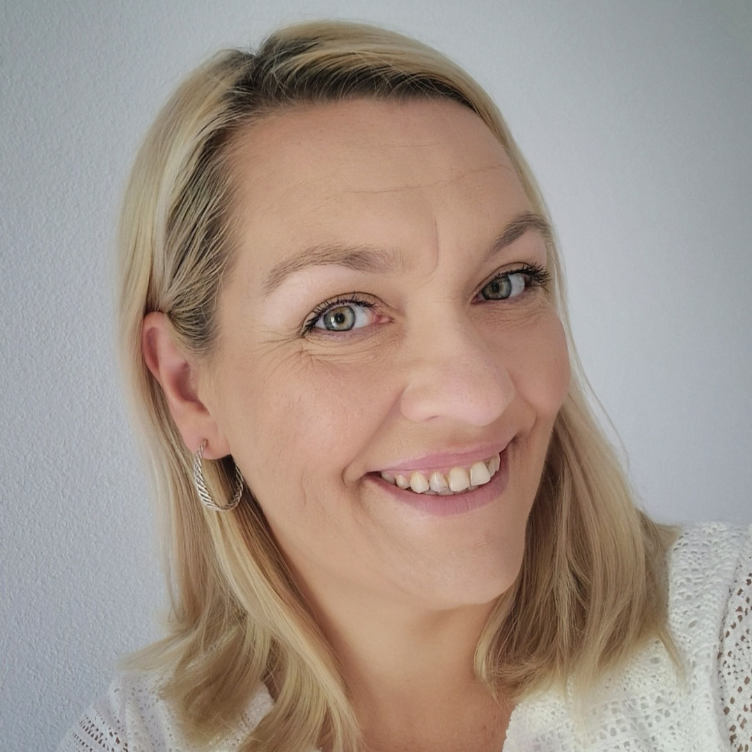 Eine Person mit blonden, schulterlangen Haaren und weißem Oberteil, die Person hält den Kopf schräg und lächelt