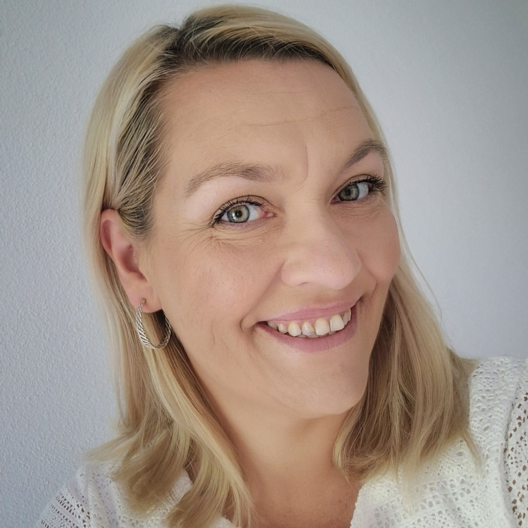 Eine Person mit blonden, schulterlangen Haaren und weißem Oberteil, die Person hält den Kopf schräg und lächelt