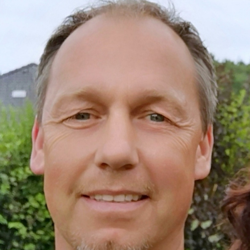 Portraitaufnahme einer Person mit kurzen Haaren und blauem Oberteil. Im Hintergrund Himmel und grüne Landschaft