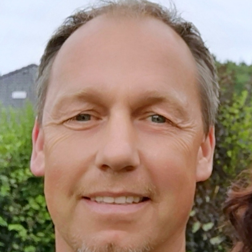 Portraitaufnahme einer Person mit kurzen Haaren und blauem Oberteil. Im Hintergrund Himmel und grüne Landschaft