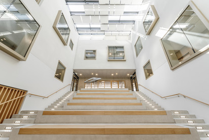 Blick in ein großes, modernes Treppenhaus. Mehrere breite Treppenläufe und Podeste verlaufen übereinander. Links sind holzfarbene Stufen zu sehen, während helle Wände und Deckenleuchten den Innenraum prägen.
