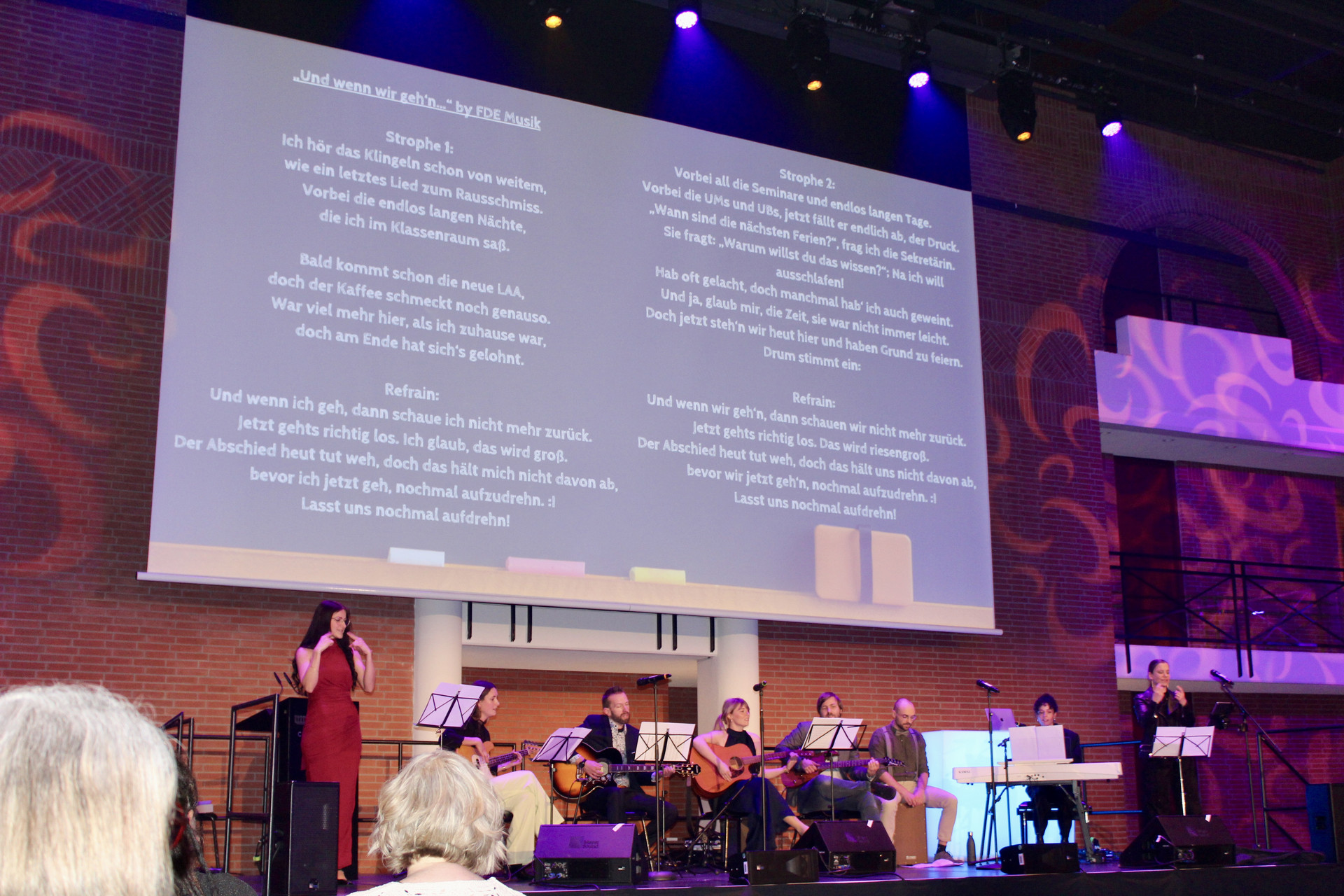 Die Gruppe der Fachdidaktischen Ergänzung Musik spielt ein Lied auf der Bühne. Im Hintergrund wird der Text eingeblendet