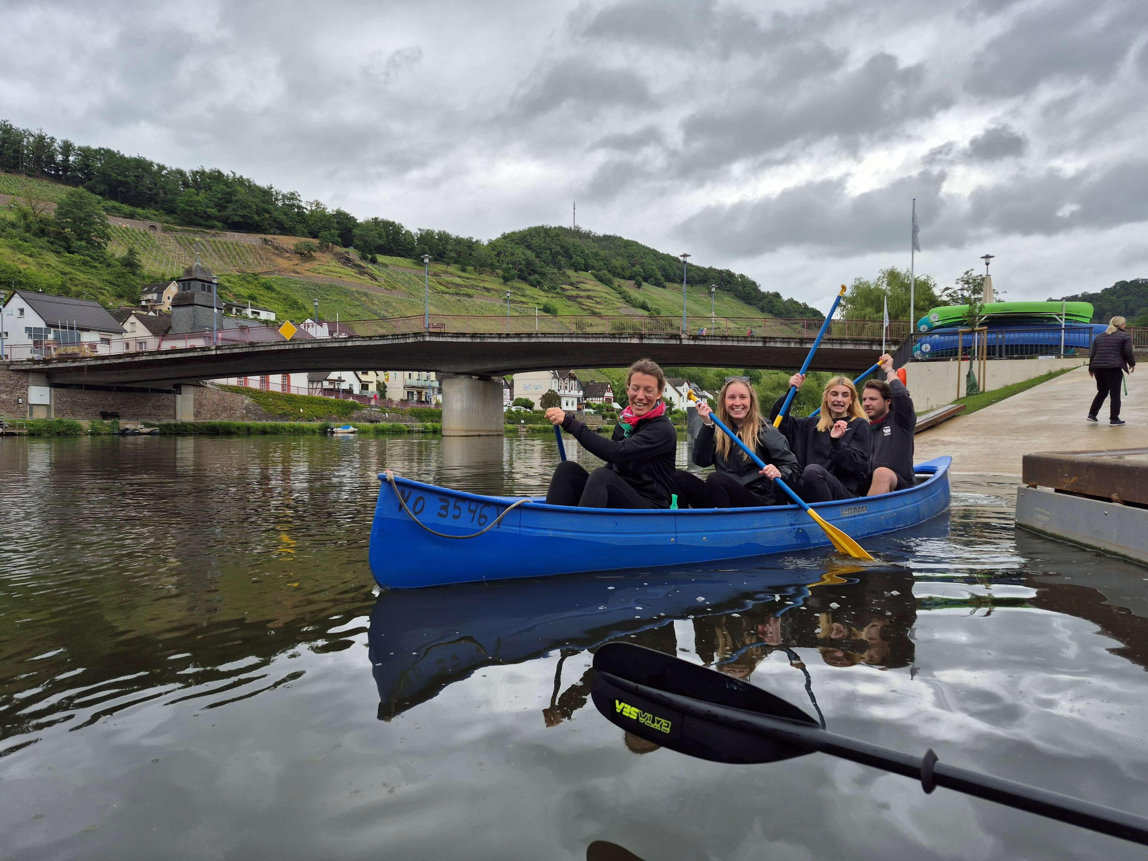 Drei Anwärterinnen und ein Anwärter sitzen in einem blauen Kanadier beim Start der Paddeltour auf der Lahn in Obernhof. Alle haben ein blau-gelbes Stechpaddel in der Hand. Im Hintergrund sieht man eine Autobrücke und dahinter Weinberge. Der Himmel in wolkenverhangen.