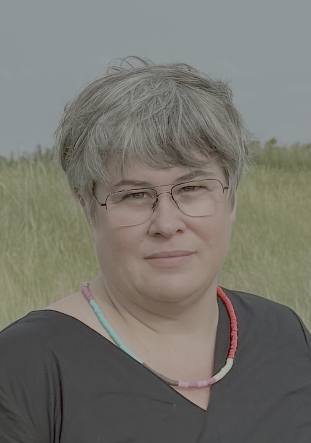 Portrait einer Person mit kurzen Haaren und Brille, schwarzem Oberteil und Kette. im Hintergrund Himmel und grüne Landscahft