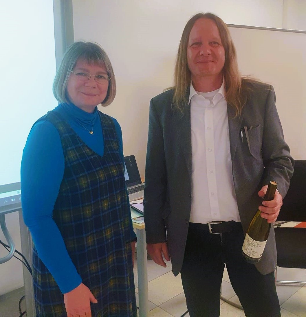 Frau Dr. Grüll vom Landesprüfungsamt führt Herrn Alexander Fähnrich in seine neue Aufgabe als Seminarleiter ein. Zur Begrüßung gibt es eine Flasche Wein aus ökologischem Anbau, die Herr Fähnrich in der Hand hält. Frau Dr. Grüll wünscht Herrn Fähnrich alles Gute für seine kommenden Aufgaben.