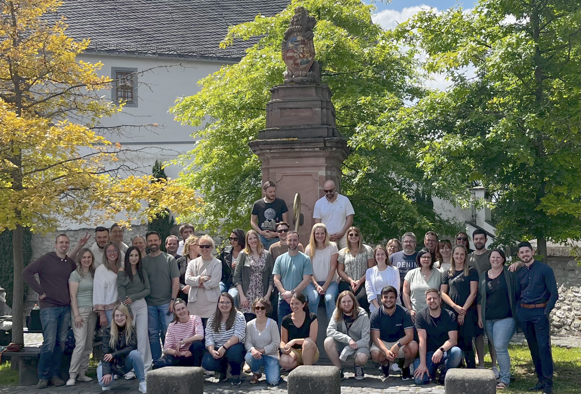 Vor einem drei Meter hohen Denkmal aus Stein, das auf einem Platz vor der Jugendherberge Diez steht, befindet sich die Gesamtgruppe der 36 Referendare und Referendarinnen sowie ihre drei Begleiter und Begleiterinnen. In der ersten Reihe knieen von links vier Referendarinnen und drei Referendare. In der zweiten Reihe stehen 14 Referendarinnen und Referendare, in der dritten Reihe 13 sowie die 3 Begleiter und Begleiterinnen. Direkt vor dem Denkmal auf einem Podest stehen in vierter Reihe 2 Referendare. Im Hintergrund sieht man drei Laubbäume, dahinter verdeckt ein Gebäude, die Sonne scheint.