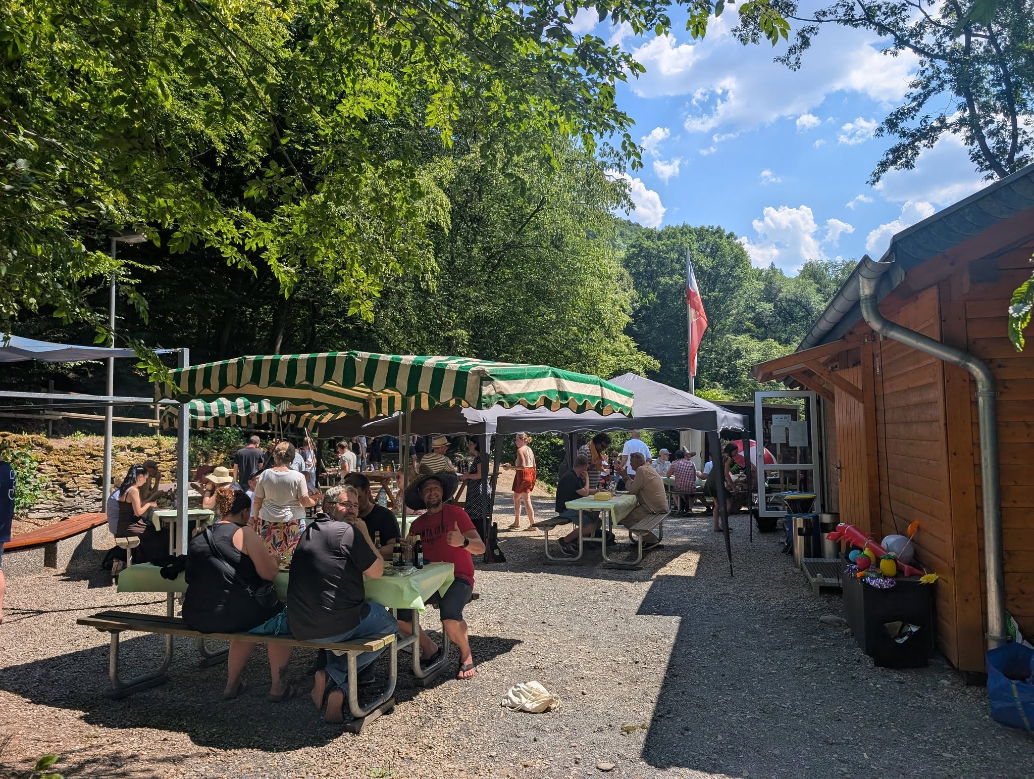 Ein Sommerfest findet auf einem sonnigen, bewaldeten Gelände mit mehreren überdachten Sitzbereichen statt. Menschen sitzen an Biertischen unter grünen und weißen Markisen sowie schwarzen Pavillons. Sie unterhalten sich, essen und trinken. Im Vordergrund sitzen Personen an einem Tisch, einer davon zeigt Daumen hoch in die Kamera. Rechts steht eine Holzhütte, daneben zwei Tonnen mit bunten Spielzeugen. Im Hintergrund weht eine Fahne, die Szenerie ist lebendig und entspannt bei blauem Himmel mit einigen Wolken.