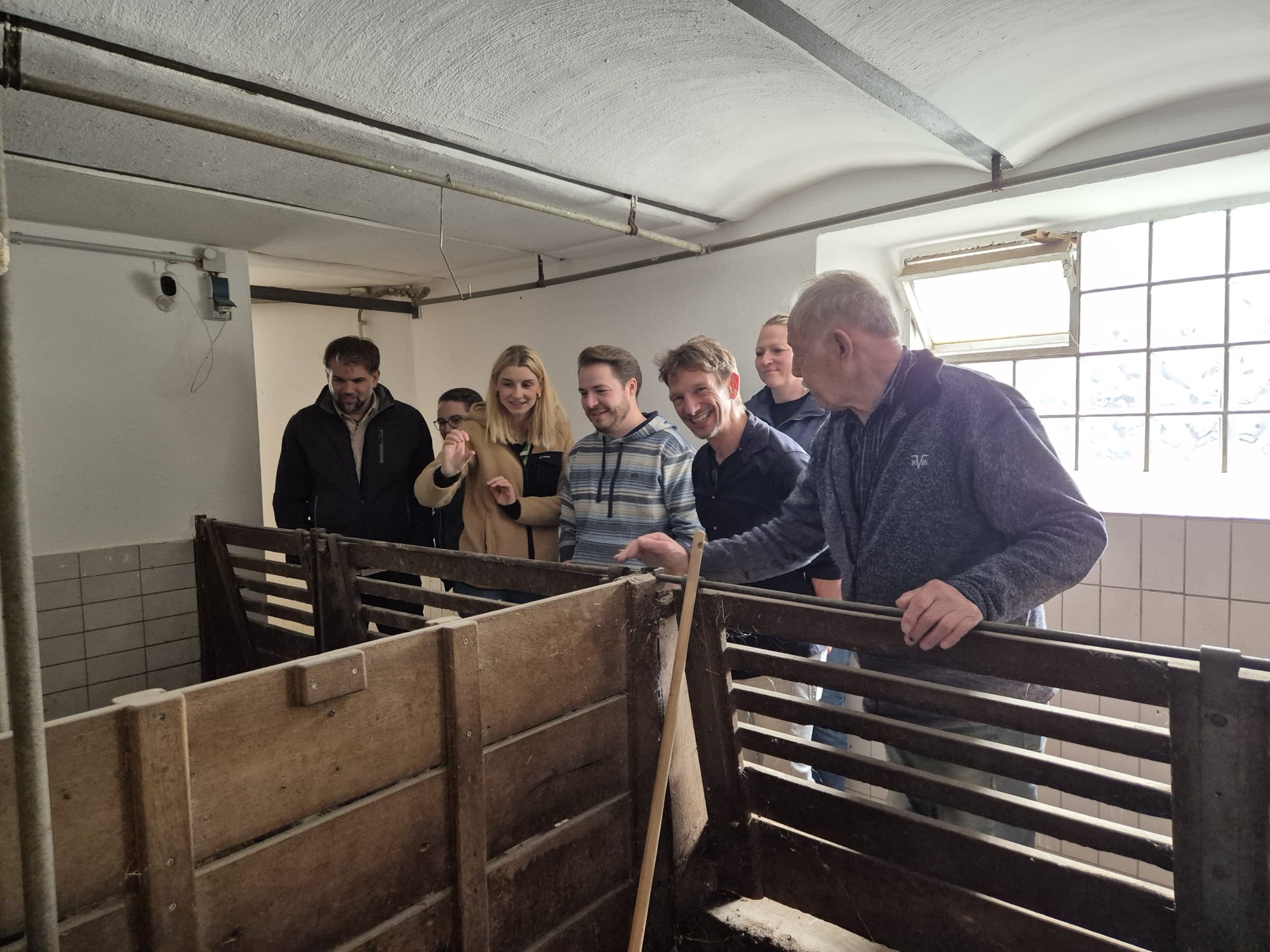 Der Weg auf dem Rundweg durch den Bauernhof führt auch durch den Schweinestall. Eine Gruppe von Personen steht vor den Schweineboxen.
