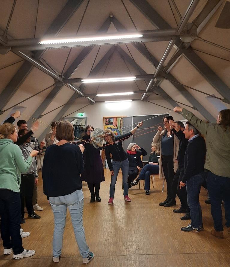 12 Teilnehmer:innen führen die Übung PerspActiv durch. Dazu stehen sie in einem Kreis und halten jeweils eine Schnur in der Hand. Am Ende der Schnüre hängt ein Wirrwarr von Schläuchen, durch die eine Kugel geführt werden soll.