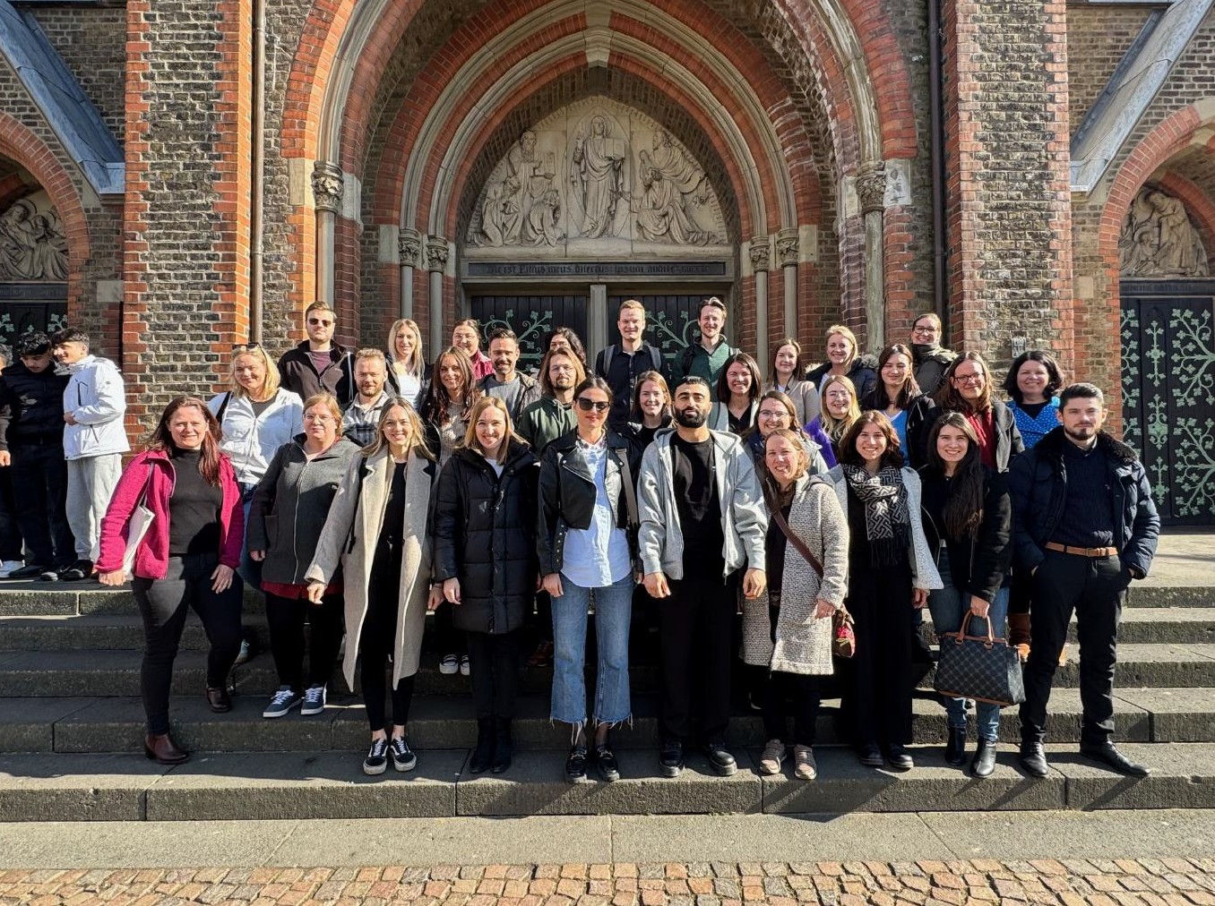 Gruppenfoto der Personen in den Fachseminaren Deutsch, Ethik und Englisch vor dem Eingang der katholischen Kirche St. Josef in Bonn. Abgebildet sind 31 gut gelaunte Personen an einem sonnigen Tag im März. Im Hintergrund ist der Eingang zur Kirche zu sehen.